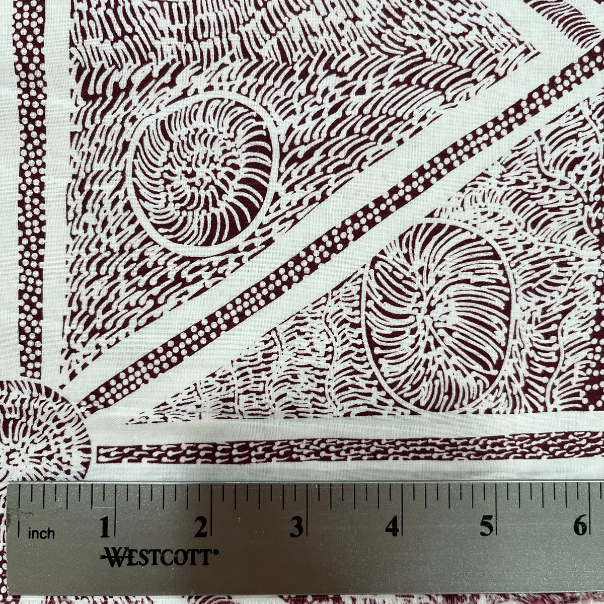A close-up of a white fabric with a dark brown geometric and organic pattern, featuring a metal ruler at the bottom showing measurements in inches. 