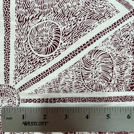 A close-up of a white fabric with a dark brown geometric and organic pattern, featuring a metal ruler at the bottom showing measurements in inches. 