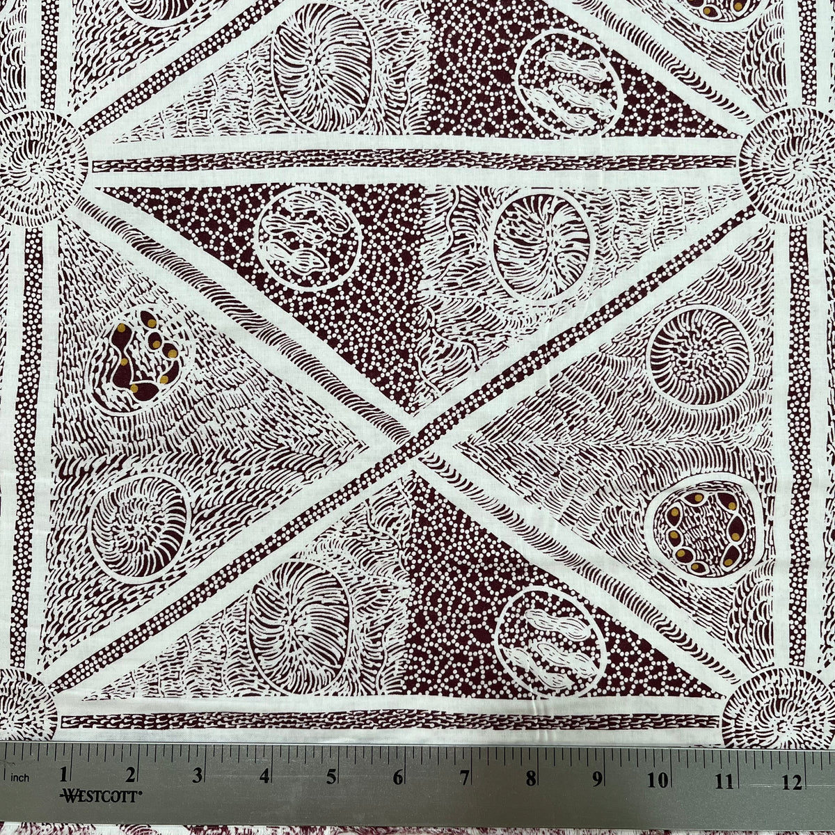 white and burgundy patterned fabric with a geometric design featuring triangles, dots, and lines, shown with a metal ruler at the bottom indicating scale.