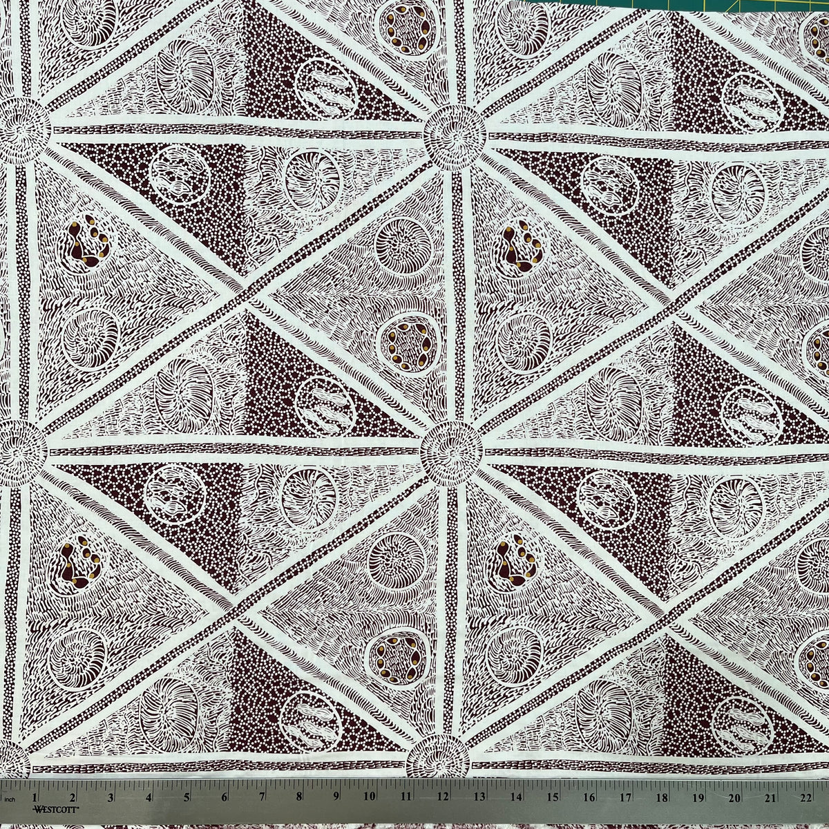 white and burgundy fabric with a repeating geometric pattern of triangles and circular medallions, shown with a metal ruler at the bottom indicating scale.