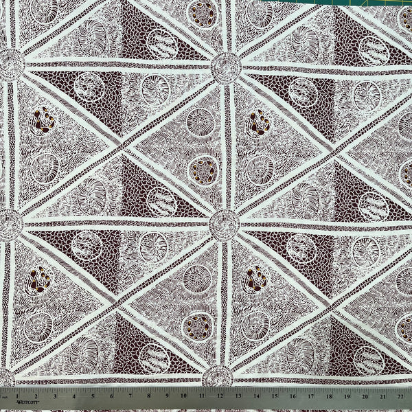 white and burgundy fabric with a repeating geometric pattern of triangles and circular medallions, shown with a metal ruler at the bottom indicating scale.