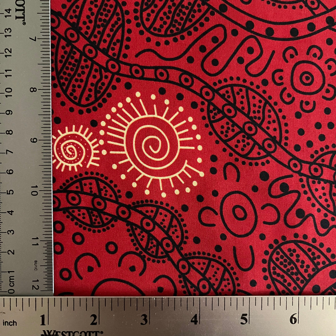 Red fabric with a black and yellow Aboriginal-style dot and spiral pattern, shown with a ruler to indicate scale.