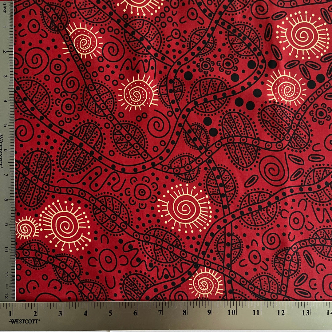 Red fabric with a black and yellow Aboriginal-style dot art pattern, shown with a metal ruler on the left side to indicate scale.