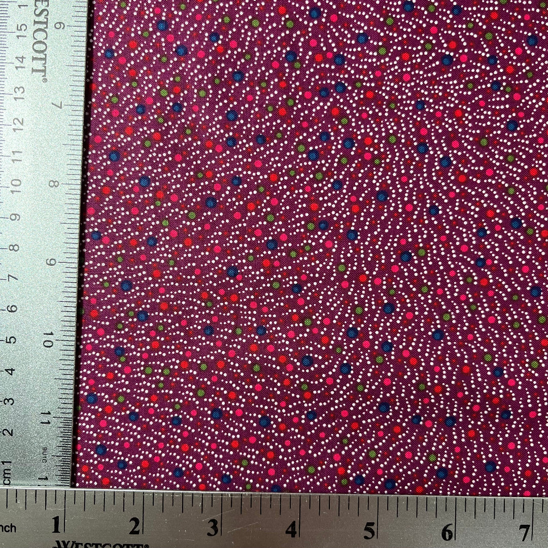 A close-up of patterned fabric with a ruler on the left and bottom edges showing measurements. The fabric has a deep magenta-purple background with dense dotted lines in white, red, pink, blue, and green.