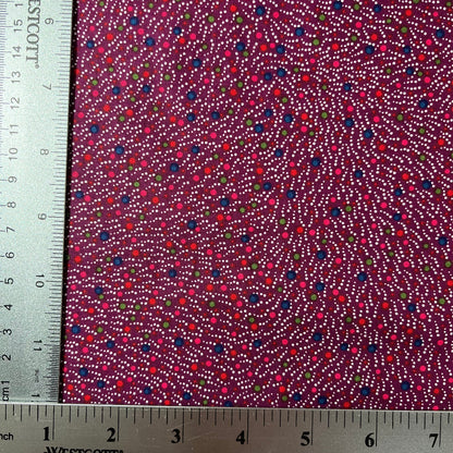 A close-up of patterned fabric with a ruler on the left and bottom edges showing measurements. The fabric has a deep magenta-purple background with dense dotted lines in white, red, pink, blue, and green.