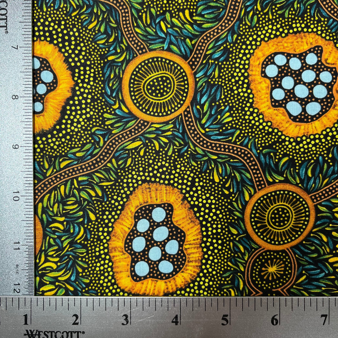 A close-up of a patterned fabric with a ruler on the left and top edges showing measurements. The fabric features a vibrant, Aboriginal dot-art style design with yellow-orange circular motifs containing light blue dots, green and yellow leafy patterns, and brown winding lines on a dark background.