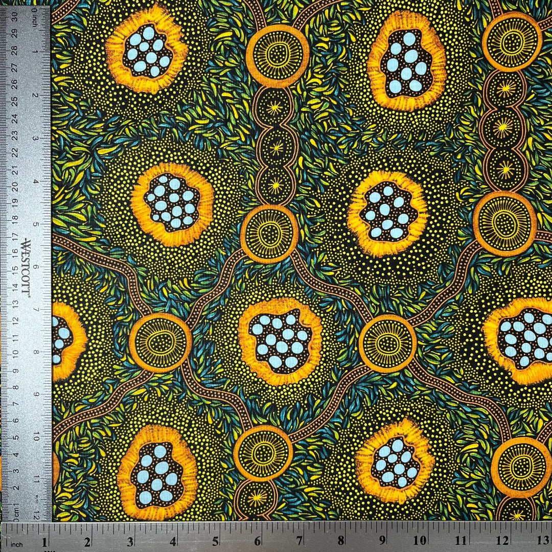 A close-up of a patterned fabric with a ruler on the left and top edges showing measurements. The fabric features a vibrant, Aboriginal dot-art style design with yellow-orange circular motifs containing light blue dots, green and yellow leafy patterns, and brown winding lines on a dark background.