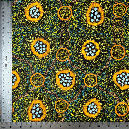 A close-up of a patterned fabric with a ruler on the left and top edges showing measurements. The fabric features a vibrant, Aboriginal dot-art style design with yellow-orange circular motifs containing light blue dots, green and yellow leafy patterns, and brown winding lines on a dark background.