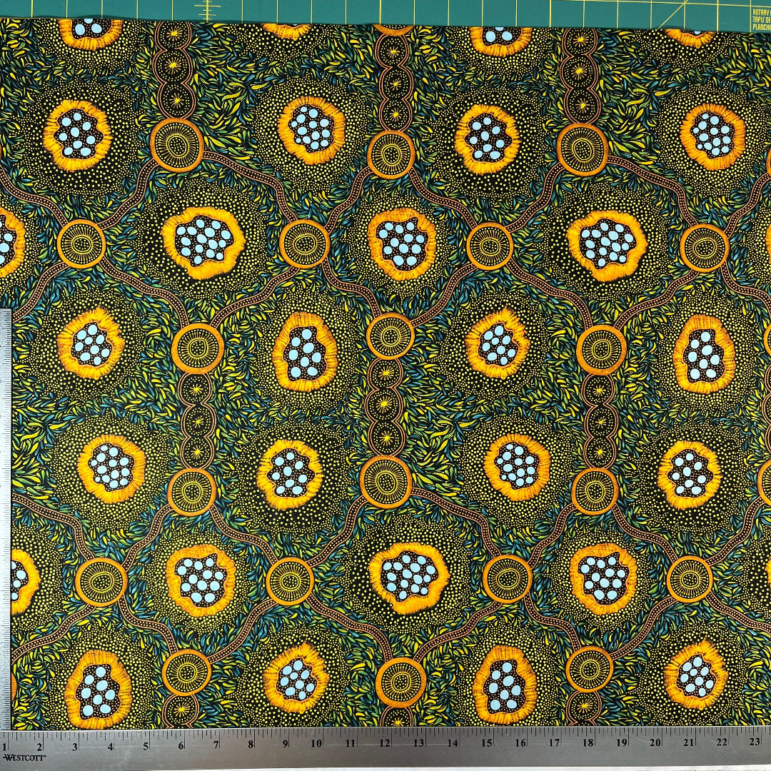 A close-up of a patterned fabric with a ruler on the left and top edges showing measurements. The fabric features a vibrant, Aboriginal dot-art style design with yellow-orange circular motifs containing light blue dots, green and yellow leafy patterns, and brown winding lines on a dark background.