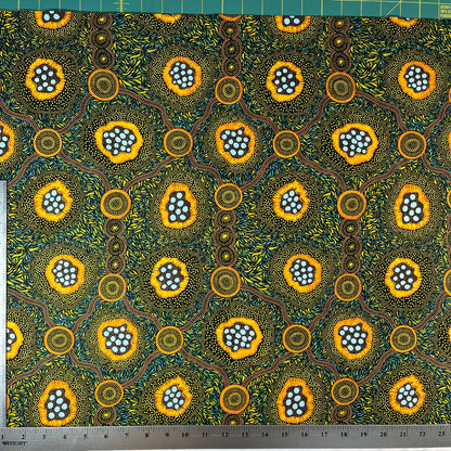 A close-up of a patterned fabric with a ruler on the left and top edges showing measurements. The fabric features a vibrant, Aboriginal dot-art style design with yellow-orange circular motifs containing light blue dots, green and yellow leafy patterns, and brown winding lines on a dark background.