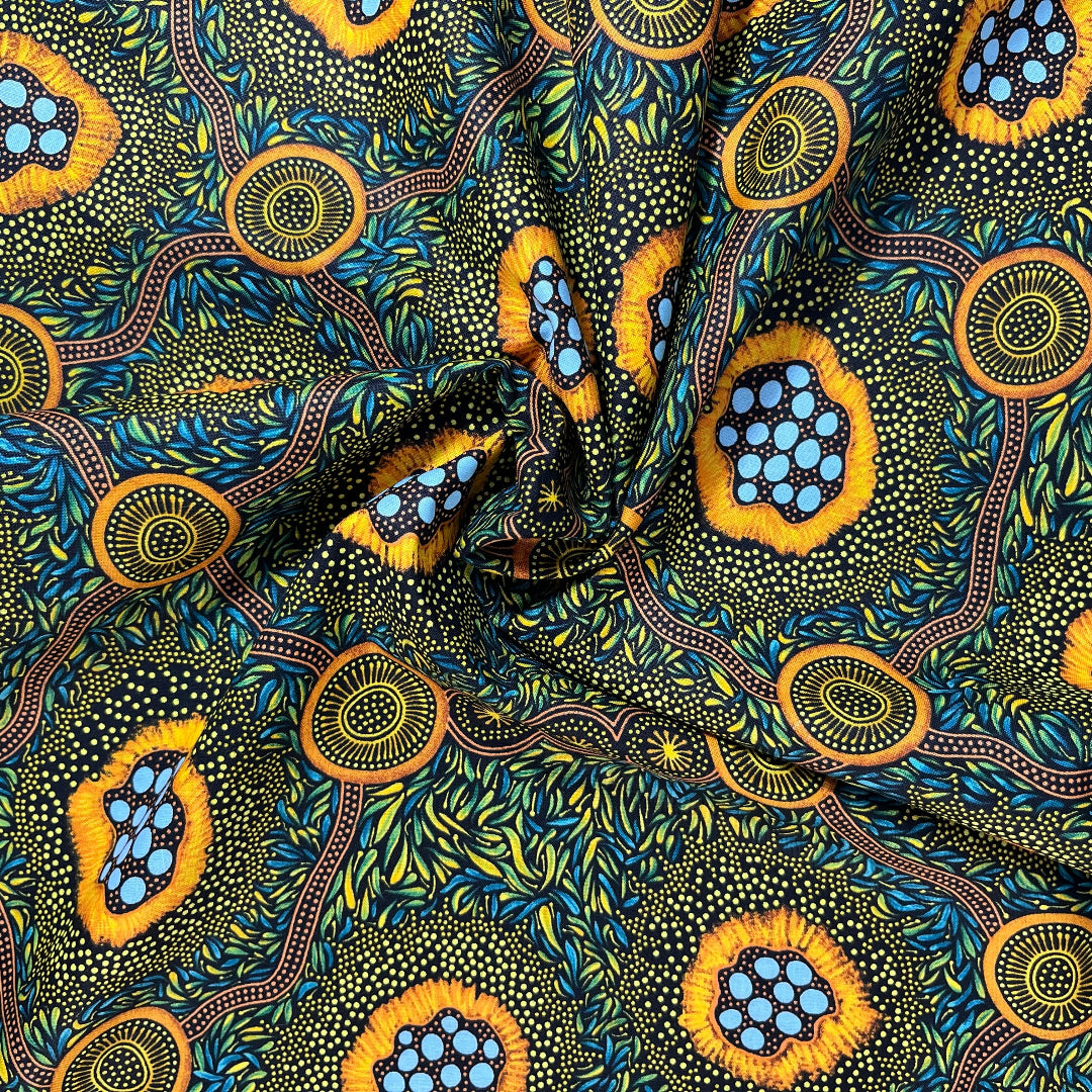 A close-up of a patterned fabric with a ruler on the left and top edges showing measurements. The fabric features a vibrant, Aboriginal dot-art style design with yellow-orange circular motifs containing light blue dots, green and yellow leafy patterns, and brown winding lines on a dark background.