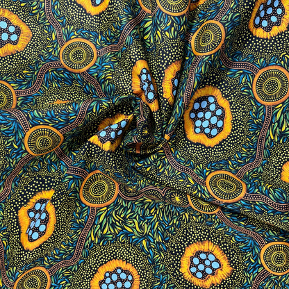 A close-up of a patterned fabric with a ruler on the left and top edges showing measurements. The fabric features a vibrant, Aboriginal dot-art style design with yellow-orange circular motifs containing light blue dots, green and yellow leafy patterns, and brown winding lines on a dark background.