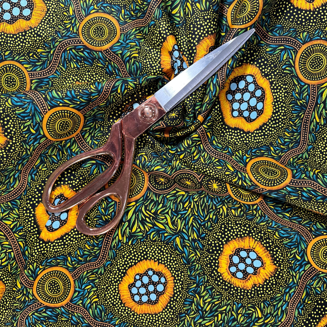 A close-up of a patterned fabric. The fabric features a vibrant, Aboriginal dot-art style design with yellow-orange circular motifs containing light blue dots, green and yellow leafy patterns, and brown winding lines on a dark background.