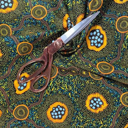 A close-up of a patterned fabric. The fabric features a vibrant, Aboriginal dot-art style design with yellow-orange circular motifs containing light blue dots, green and yellow leafy patterns, and brown winding lines on a dark background.