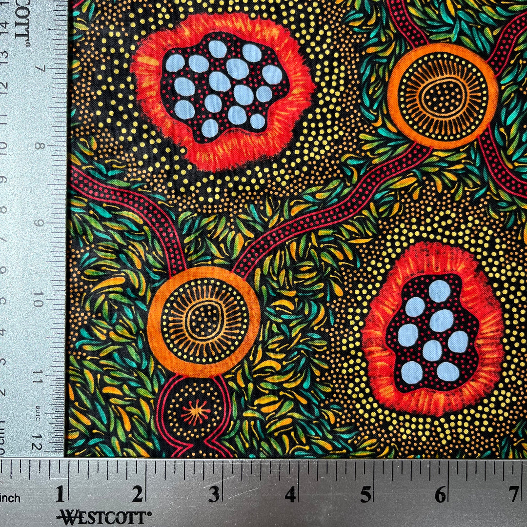A close-up of patterned fabric with a ruler along the top and left edges for scale. The fabric has a black background covered in dense yellow and green leaf-like dots, with three large circular floral or abstract medallion motifs in red, orange, and light blue.