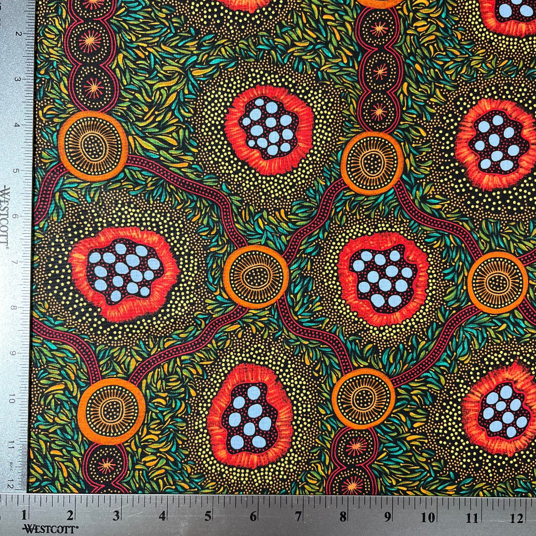 A close-up of patterned fabric with a ruler along the top and left edges for scale. The fabric has a black background covered in dense yellow and green leaf-like dots, with three large circular floral or abstract medallion motifs in red, orange, and light blue.