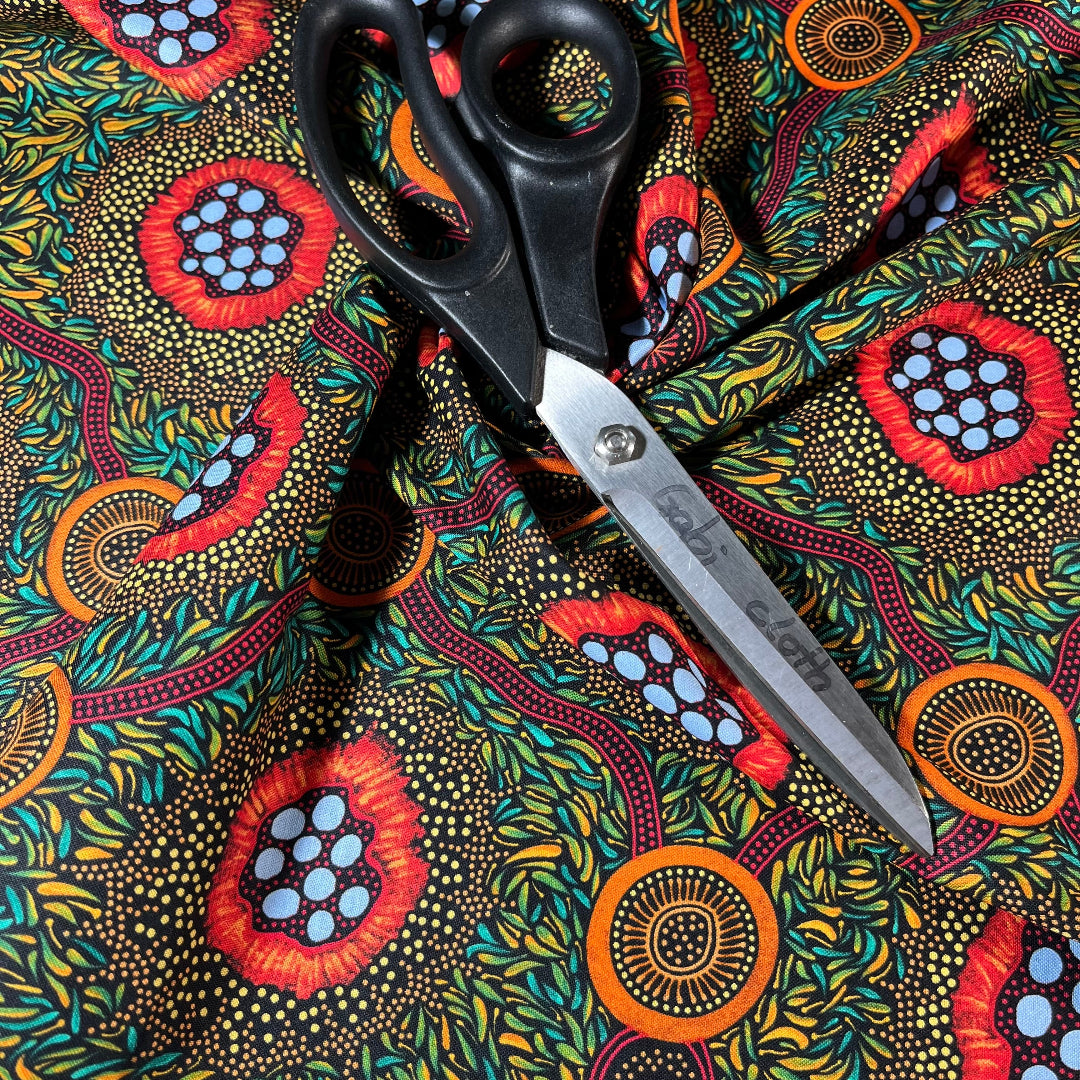 A pair of black-handled scissors with silver blades rests diagonally on a vibrant, patterned fabric. The fabric has a black background covered in dense yellow and green leaf-like dots, with three large circular floral or abstract medallion motifs in red, orange, and light blue.