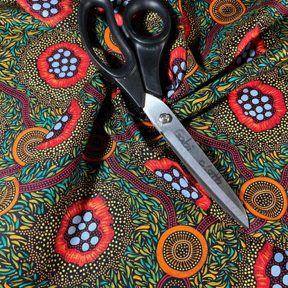 A pair of black-handled scissors with silver blades rests diagonally on a vibrant, patterned fabric. The fabric has a black background covered in dense yellow and green leaf-like dots, with three large circular floral or abstract medallion motifs in red, orange, and light blue.